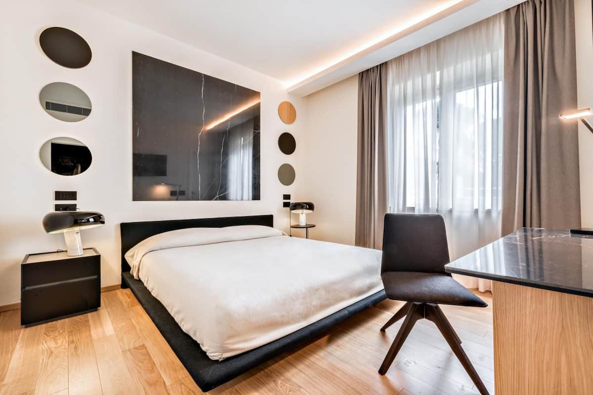 A modern hotel room with a large bed, a desk, and a chair. There are multiple wall-mounted lights and a large mirror.
