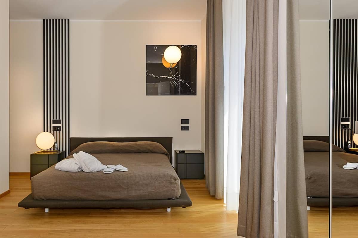 A modern hotel room with a large bed, a bedside table with a lamp, a small cabinet, and a wall-mounted artwork.
