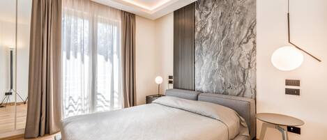 A modern bedroom with a large bed, a marble accent wall, a bedside table with a lamp, and a view through a window with curtains.