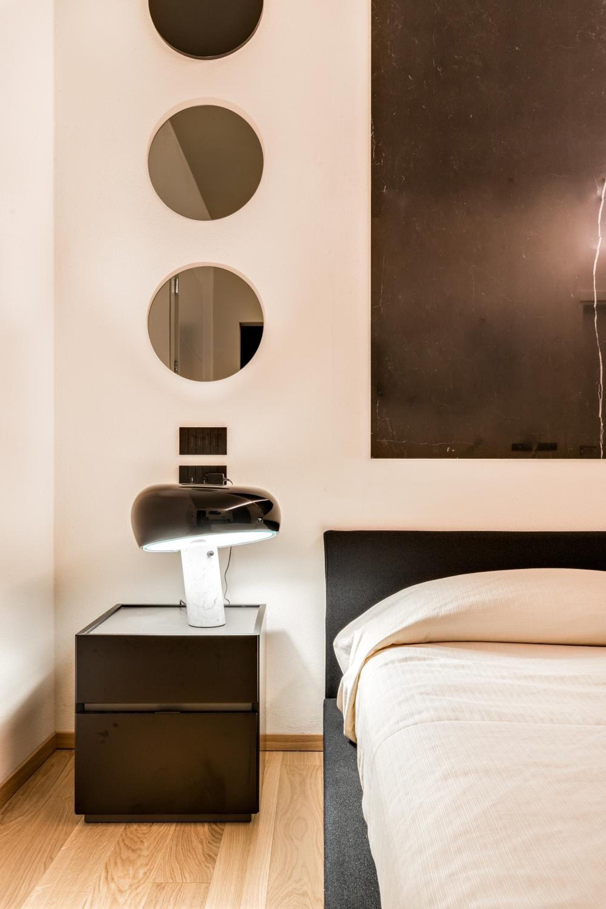 Een moderne slaapkamer met een bed, een nachtkastje met een lamp en drie spiegels aan de muur.