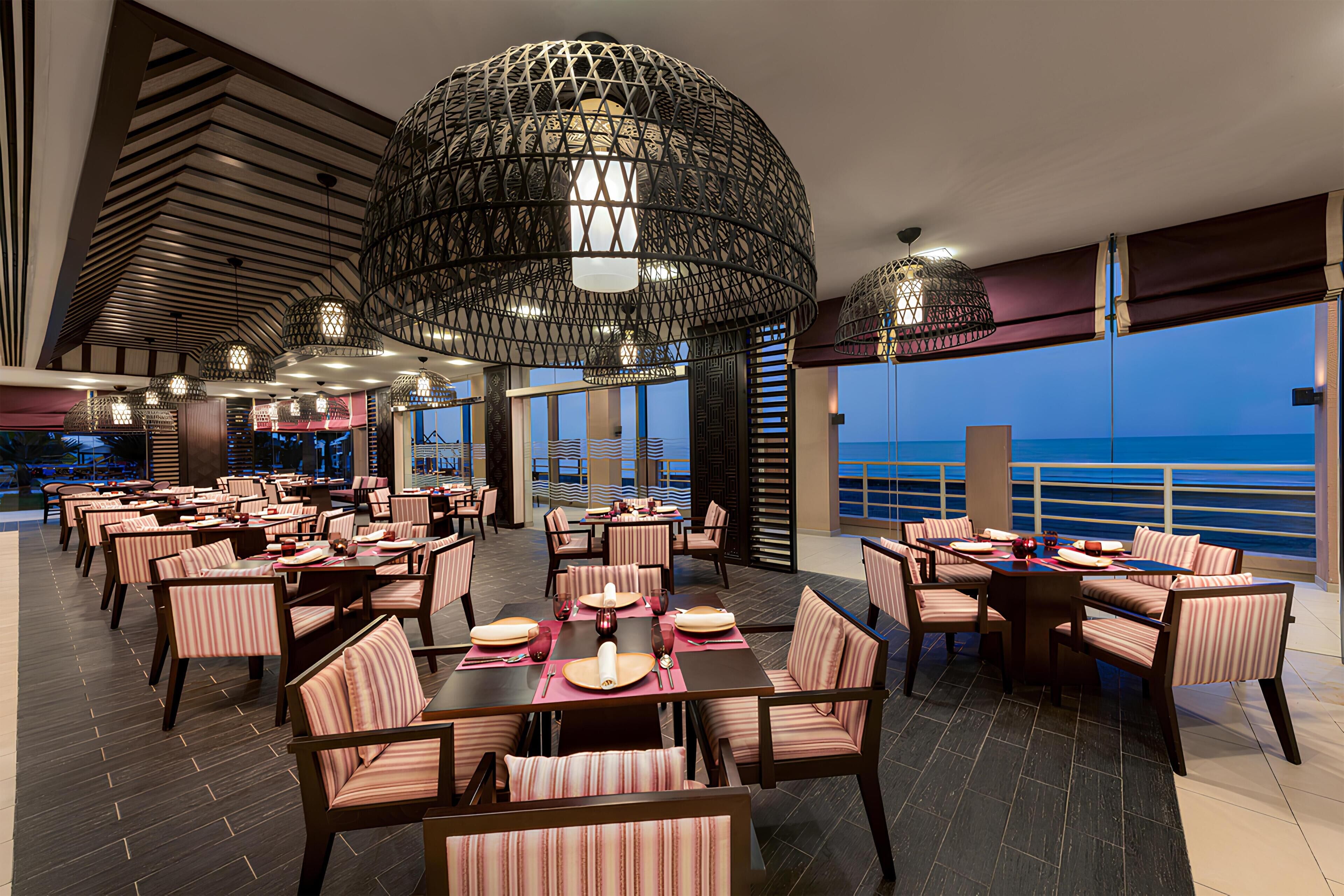 A spacious dining area with multiple tables and chairs, large hanging light fixtures, and a view of the ocean.