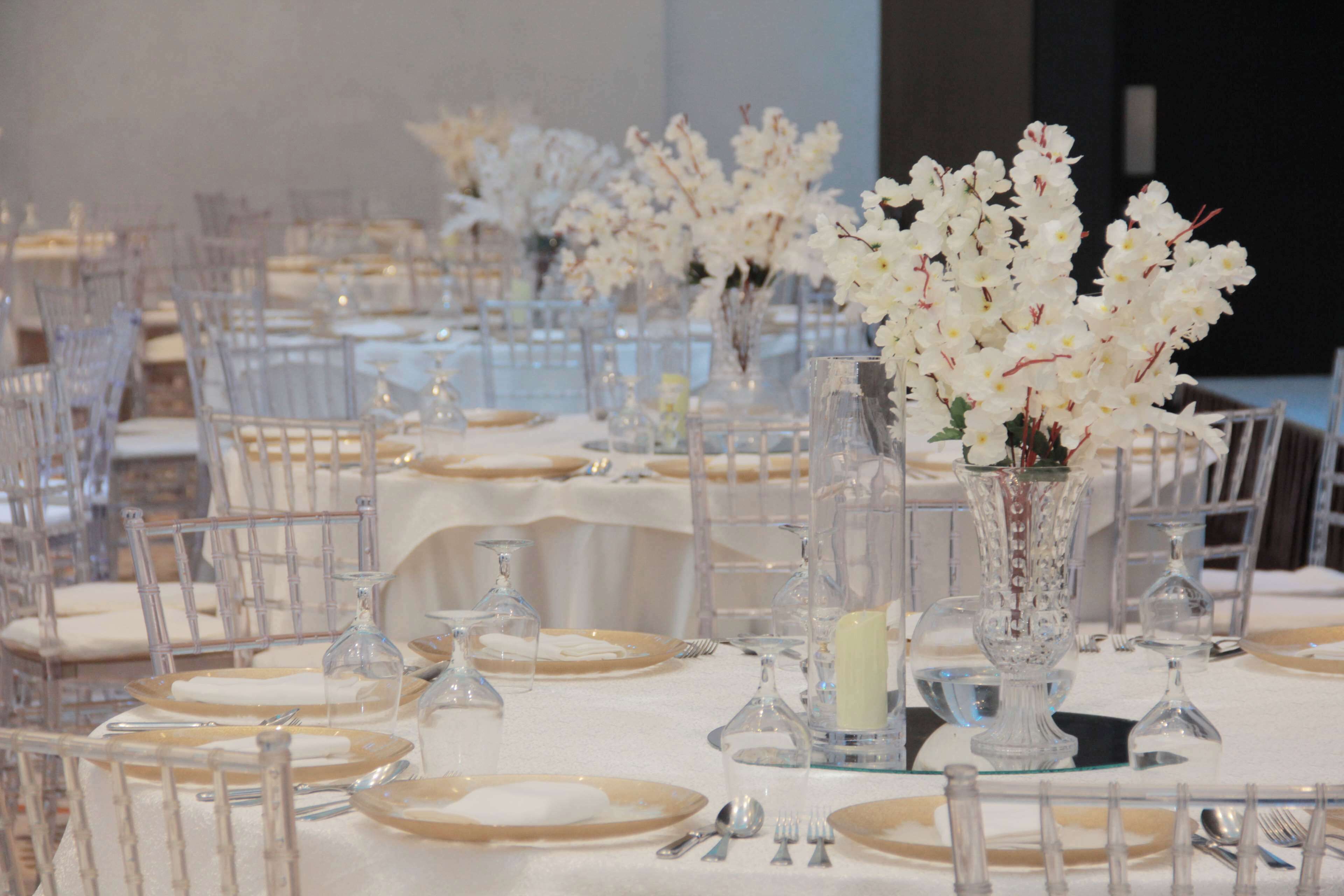 Un salón de banquetes con mesas dispuestas para un evento formal, que incluye copas de cristal, manteles blancos y centros de mesa florales.