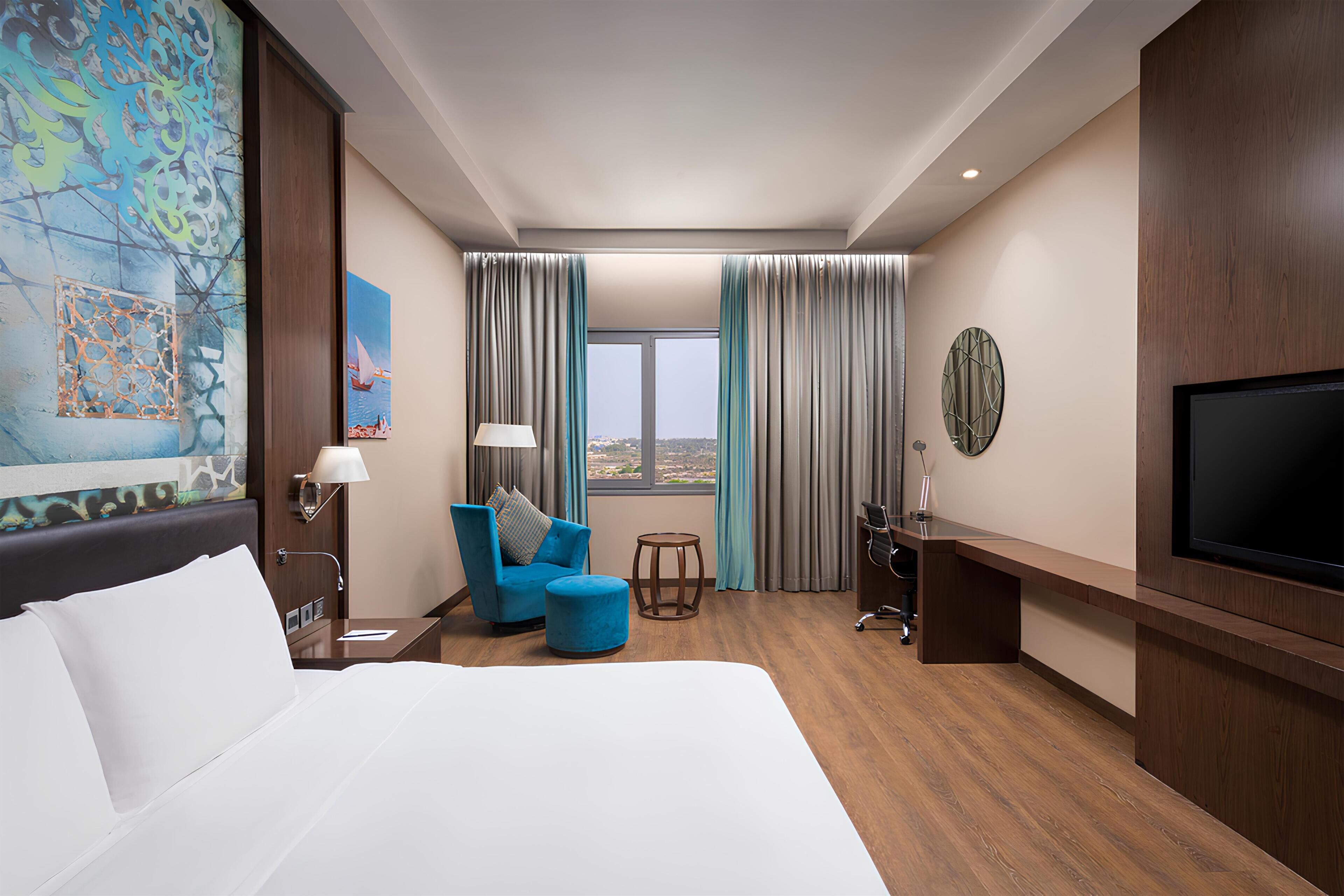 Una habitación de hotel con una cama, un sillón azul, una mesita, un televisor y una ventana con cortinas.