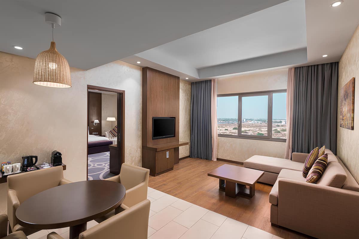 Habitación de hotel con comedor, sofá, televisión y un amplio ventanal con vistas.