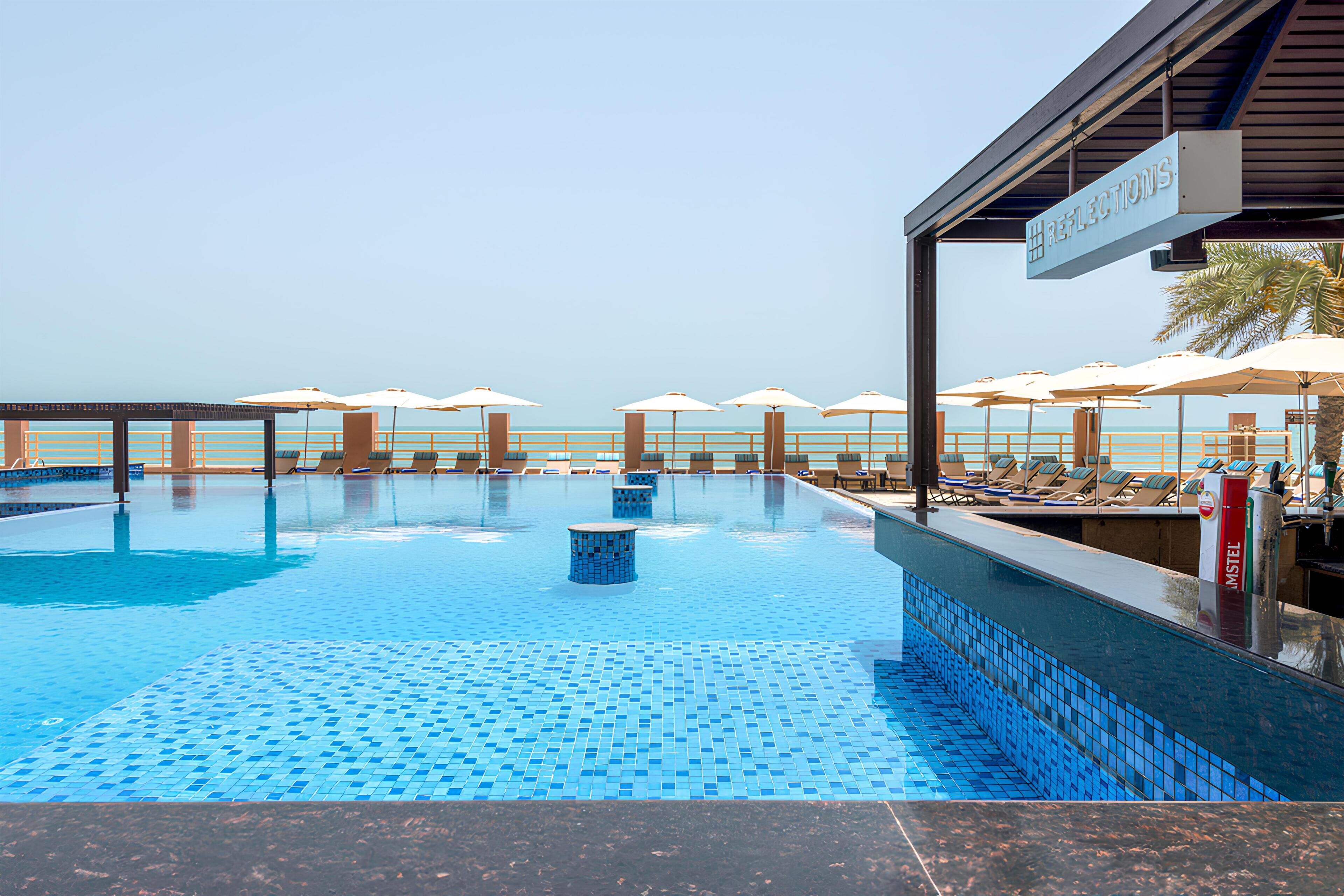 A swimming pool with lounge chairs and umbrellas, a sign for 'Reflections', and a view of the sea.