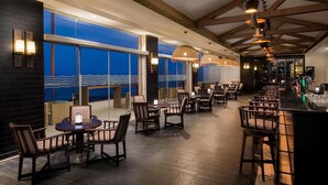 Un restaurante con barra, mesas, sillas y ventanas amplias con vistas.