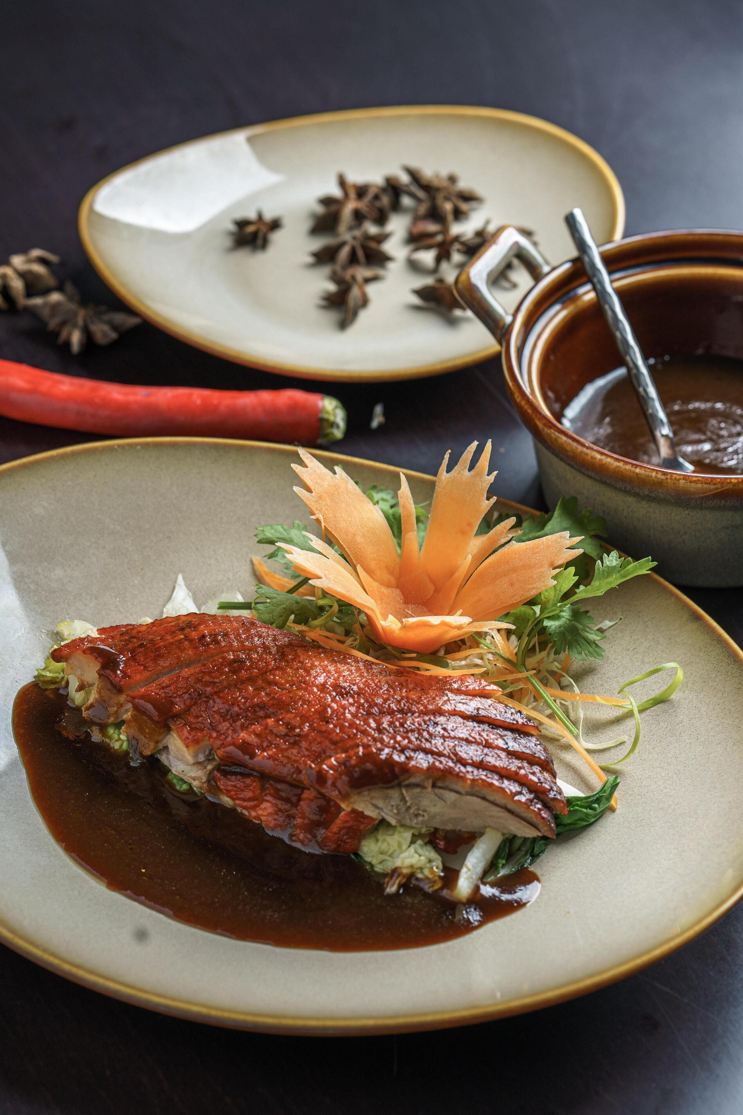 Un plato con pato asado, adornado con vegetales y una hoja decorativa, acompañado de un cuenco de salsa y anís estrellado.