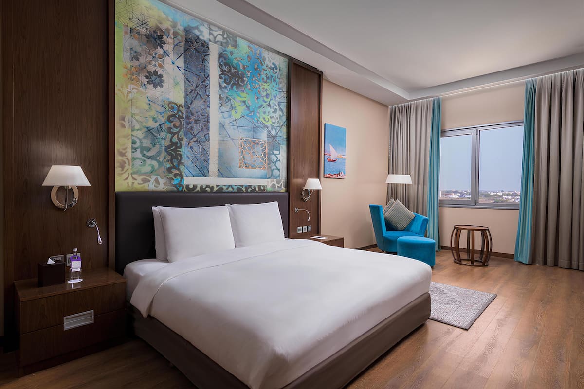 Una habitación de hotel con una cama grande, un escritorio, una silla, una ventana con cortinas y un cuadro en la pared.