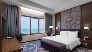 Habitación de hotel con una cama grande, un escritorio, una silla, un ventanal con vista al mar y un panel decorativo en la pared.