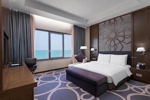 A hotel room with a large bed, a desk, a chair, a window with a view of the sea, and a decorative wall panel. - Radisson Blu Hotel & Resort, Sohar (Sohar)