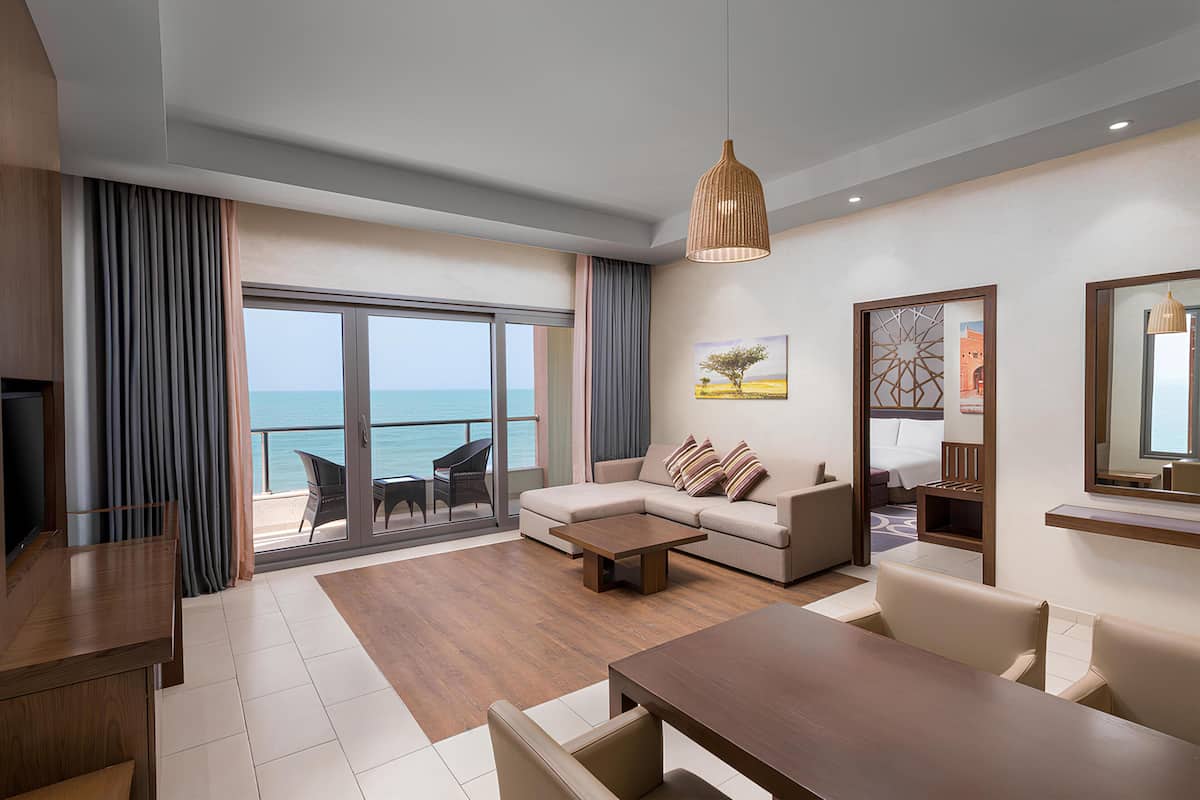 Una sala de estar moderna con sofá, mesa de centro y mesa de comedor, un televisor, y un balcón con vistas al mar.