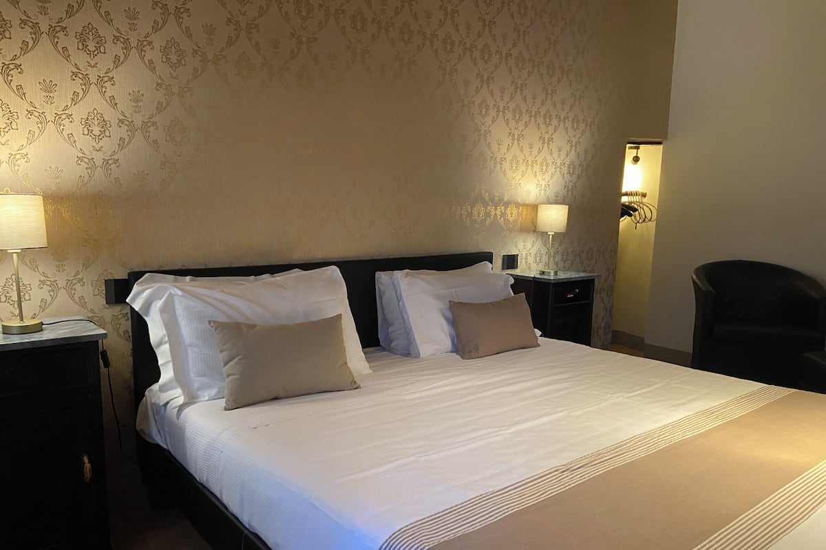 Een net opgemaakt bed met witte lakens en twee kussens, zijkanten omlijst door twee nachtkastjes met lampen, in een kamer met behang met patroon en een stoel in de hoek.