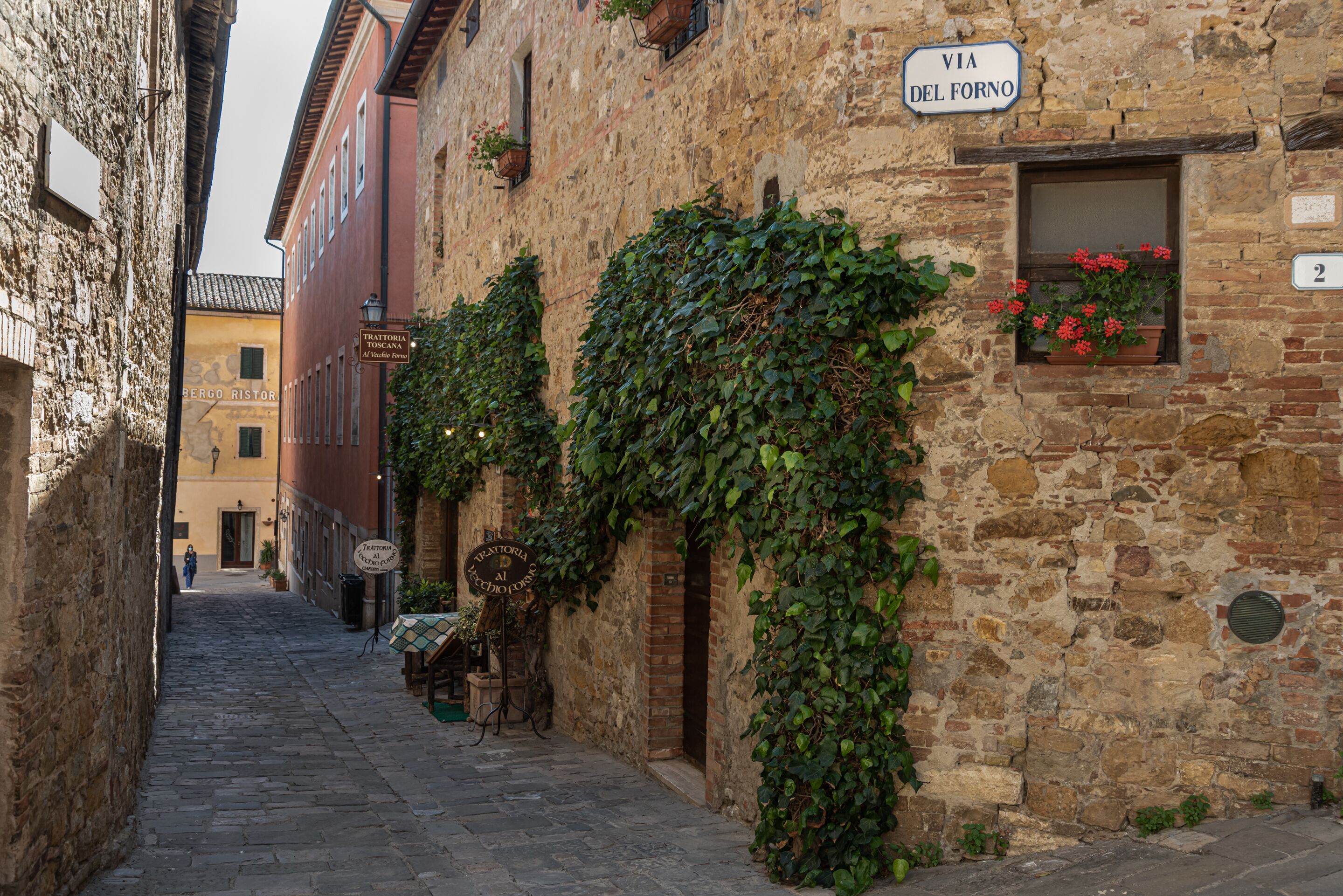 A narrow cobblestone street with stone buildings, a sign reading "VIA DEL FORNO", and a window with red flowers.