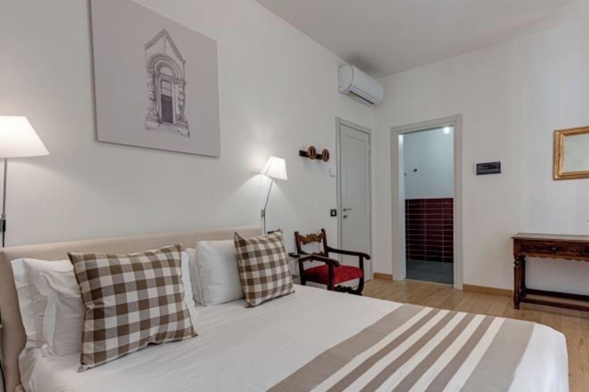 A hotel room with a bed, a chair, a table, and a door.