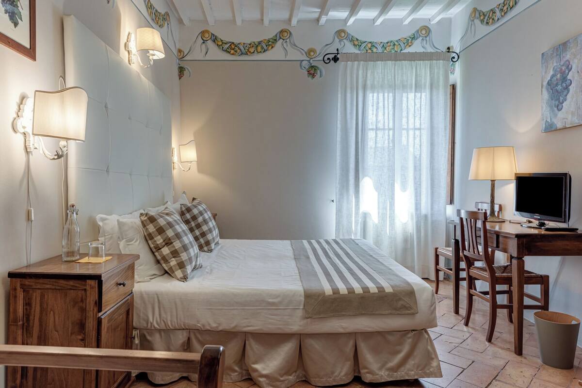 Een slaapkamer met een bed, nachtkastjes, een bureau met stoel, een televisie en een raam met gordijnen.