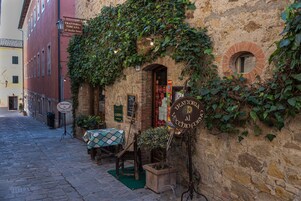 Una estrecha calle adoquinada con un edificio rústico que presenta un arco de ladrillo y paredes cubiertas de hiedra. Hay una silla de madera y una mesa con mantel a cuadros afuera.