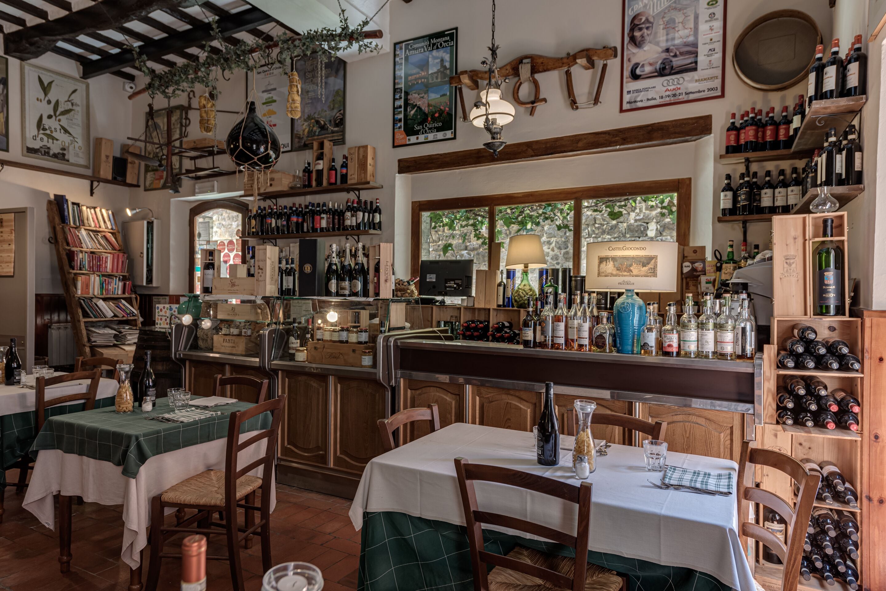 A restaurant with wooden tables and chairs, a bar area with wine bottles, and a view of trees through the windows.