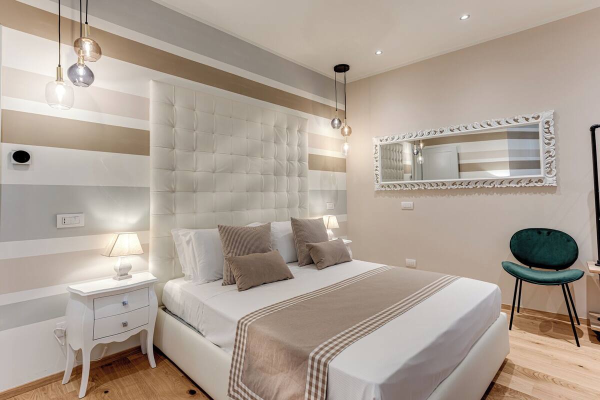 Een moderne slaapkamer met een gestoffeerd hoofdbord, een grote spiegel, een nachtkastje met een lamp en een stoel.