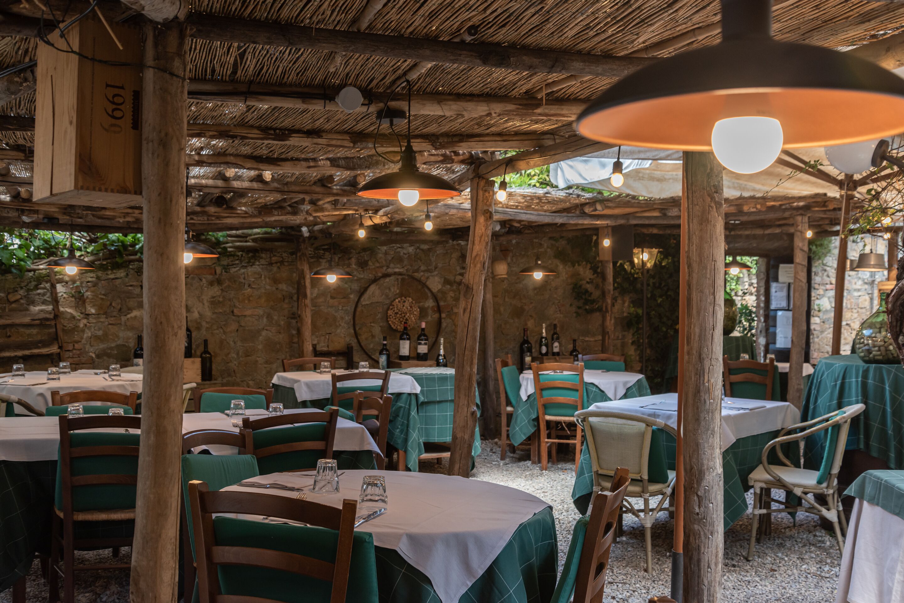 Een openluchtrestaurant met ronde tafels, groen geruit tafelkleed, houten stoelen en hangende lampen.