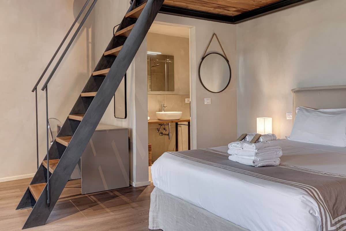 Een hotelkamer met een modern design, met een bed met netjes gevouwen handdoeken, een houten trap, een ronde spiegel en een badkamer met een wastafel en spiegel.