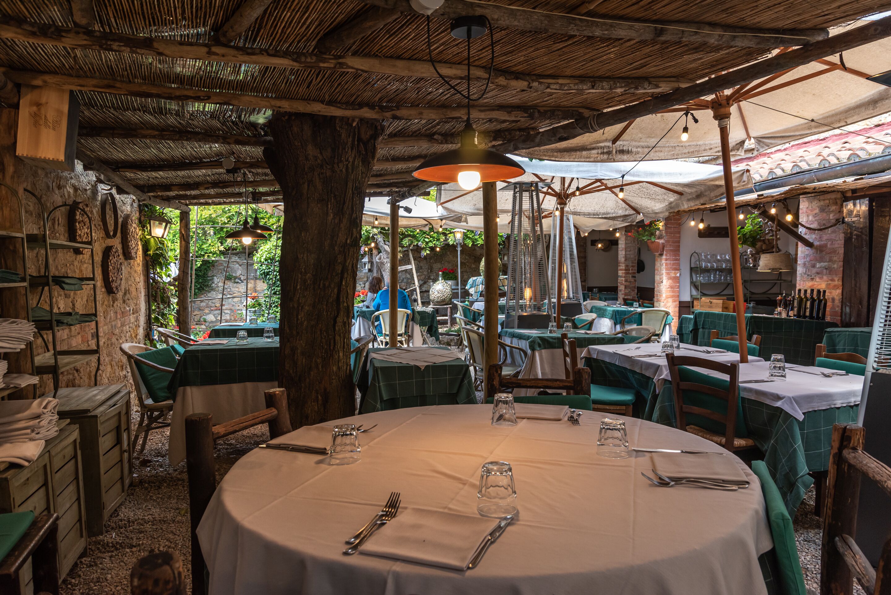 Ein Restaurant im Freien mit runden Tischen, grünen Polstersesseln und einer rustikalen Holzbalkendecke.