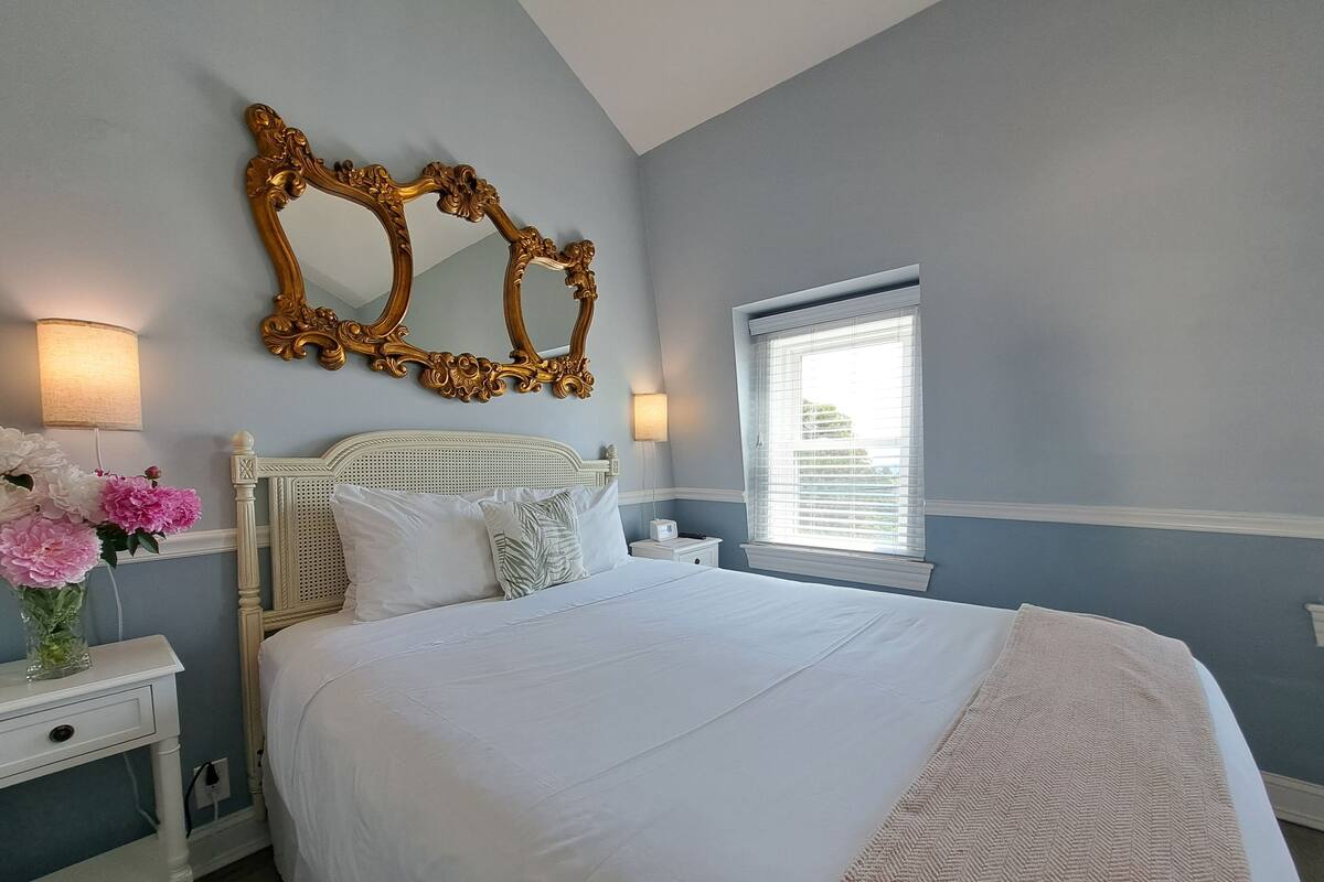 Quarto com cama grande, roupa de cama branca, espelho decorativo e mesa de cabeceira com um vaso de flores.