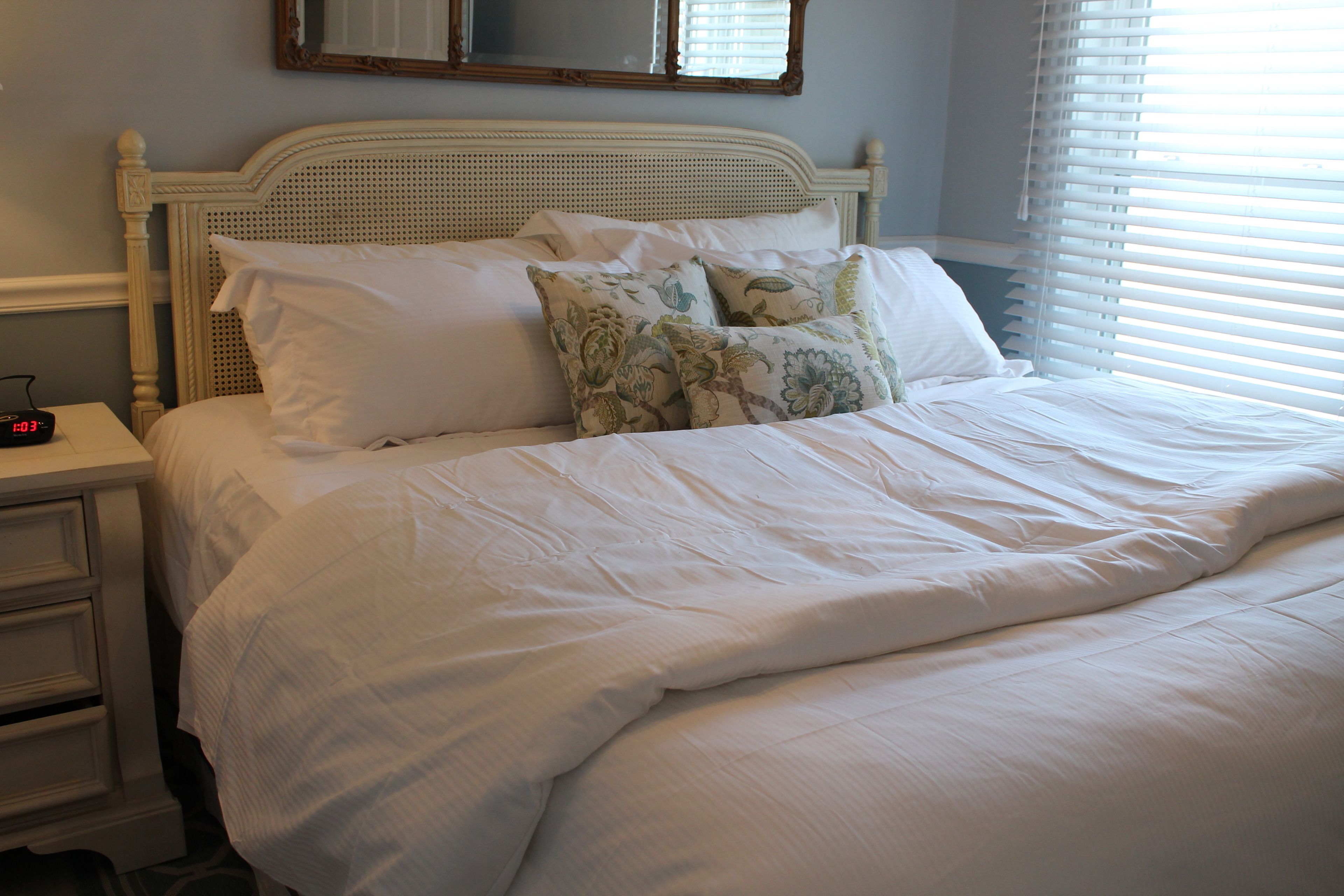A neatly made bed with white linens and floral pillows, a bedside table with a clock, and a window with blinds.