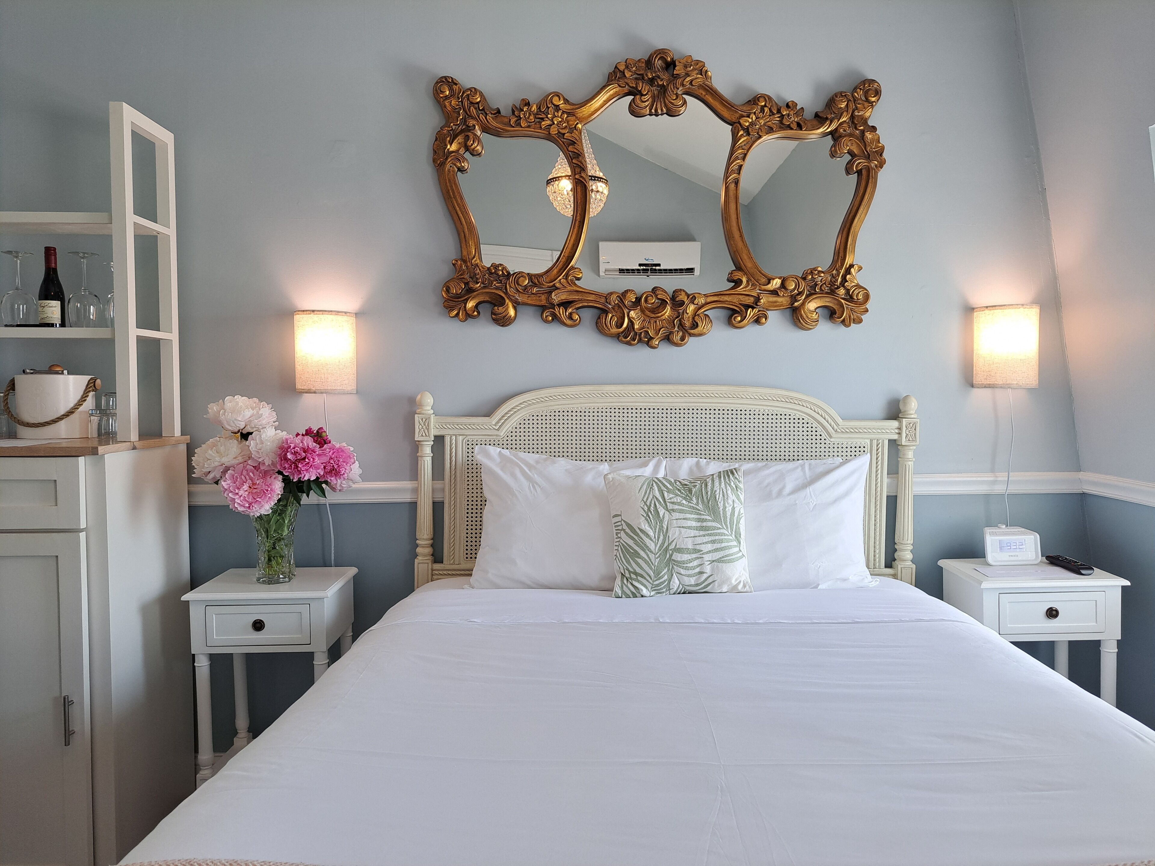 A hotel room with a large ornate mirror, a bed with white linens, a nightstand with a lamp, and a vase with flowers.