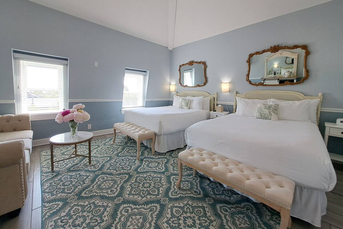 Quarto com duas camas, um banquinho, uma pequena mesa com flores e dois espelhos fixados na parede.