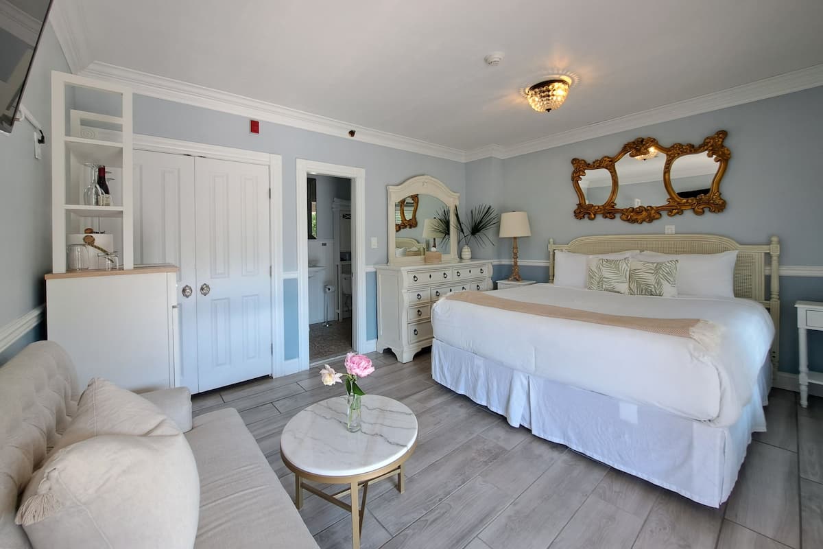 Quarto de hotel com uma cama grande, área de estar com mesa redonda, cômoda e espelho.