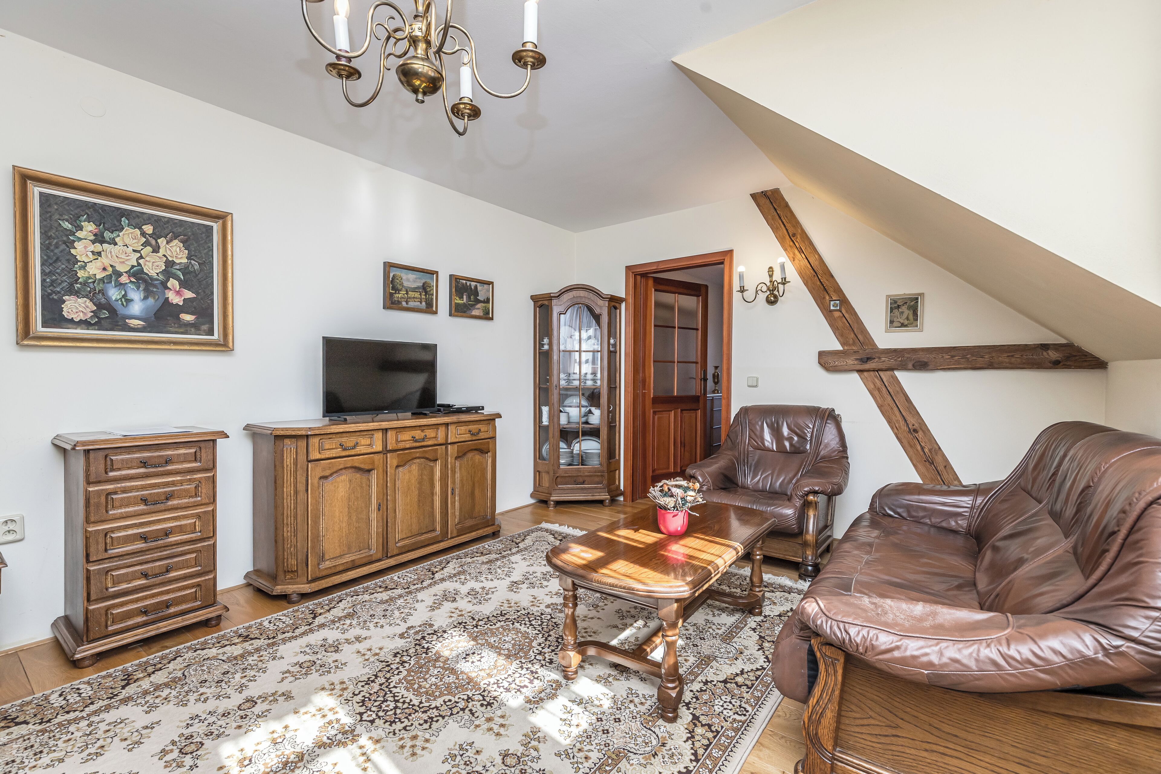 A living room with a brown leather sofa, wooden coffee table, and a TV on a wooden cabinet. There is a patterned rug on the floor, a chandelier, and a wooden door.
