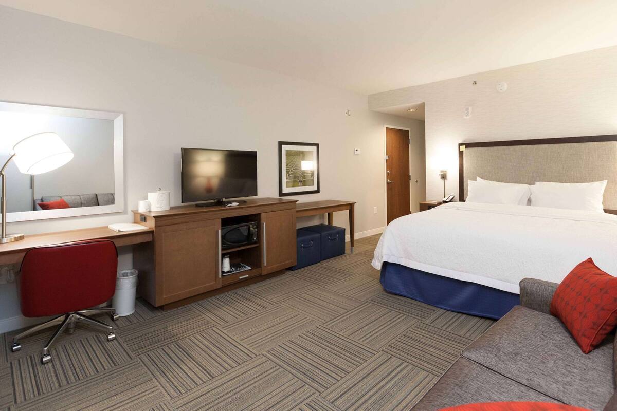 A hotel room with a bed, a desk with a TV, a chair, a microwave, and a red couch.