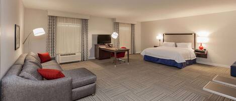 A hotel room with a sofa, a bed, a desk with a TV, and a lamp.