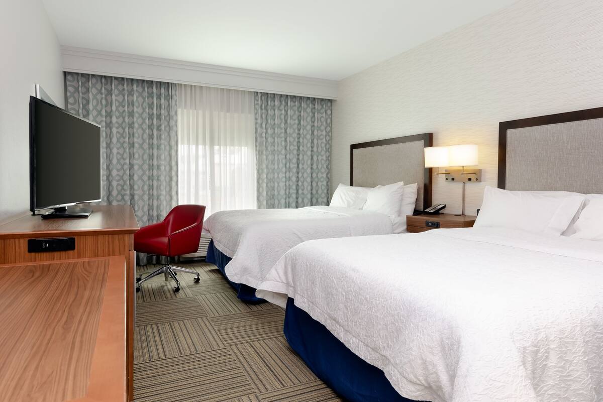 A hotel room with two beds, a desk with a TV, a red chair, and a lamp.
