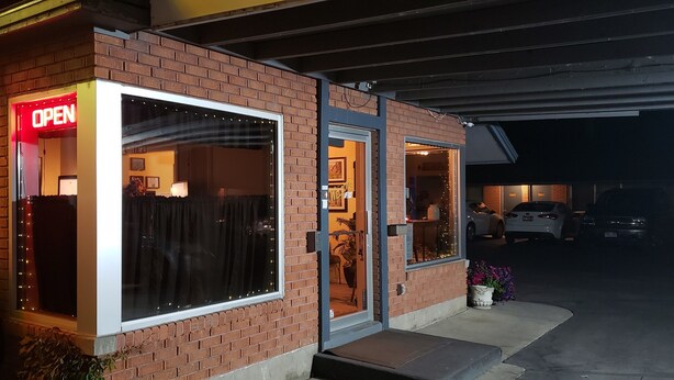 A brick building with a large illuminated 'OPEN' sign, a glass door entrance, and a small outdoor area with potted plants. - Lava Spa Motel & RV Park (Lava Hot Springs)