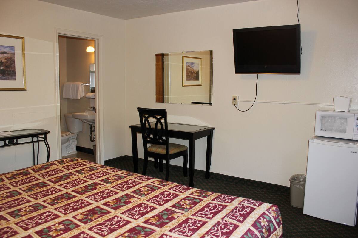 A hotel room with a bed, a desk, a chair, a television, a microwave, a trash can, and a bathroom with a toilet and sink.