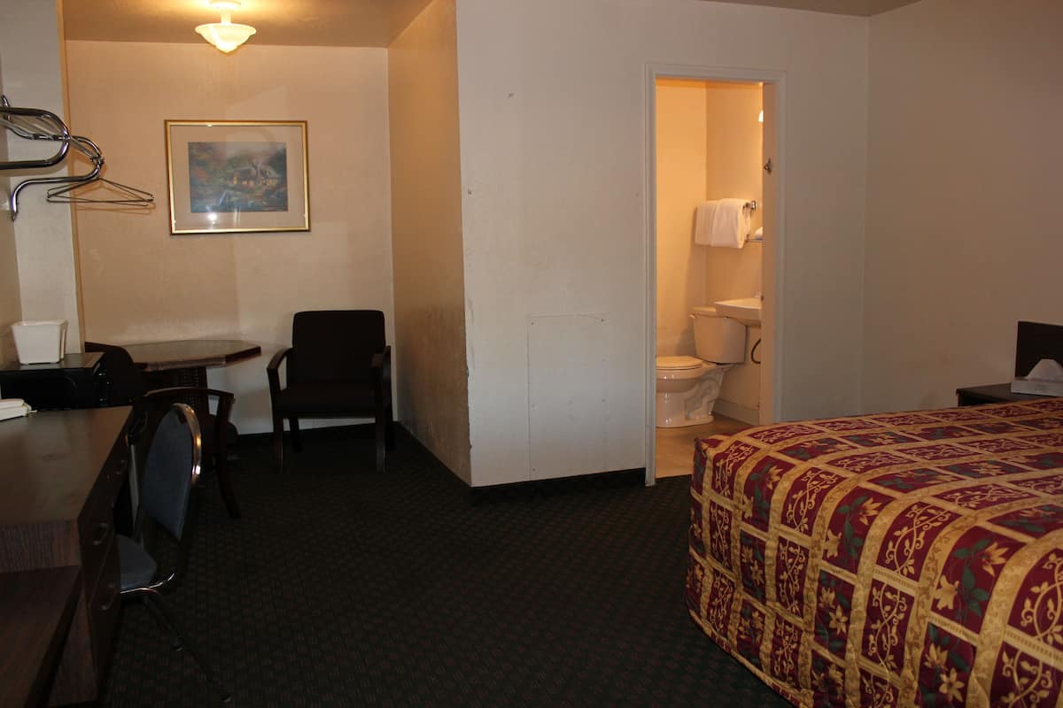 A hotel room with a bed, a desk, a chair, a small table, a framed picture on the wall, and a bathroom visible through an open door.