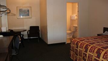 Habitación de hotel con cama, escritorio, silla, mesita, un cuadro en la pared y baño visible a través de una puerta abierta.