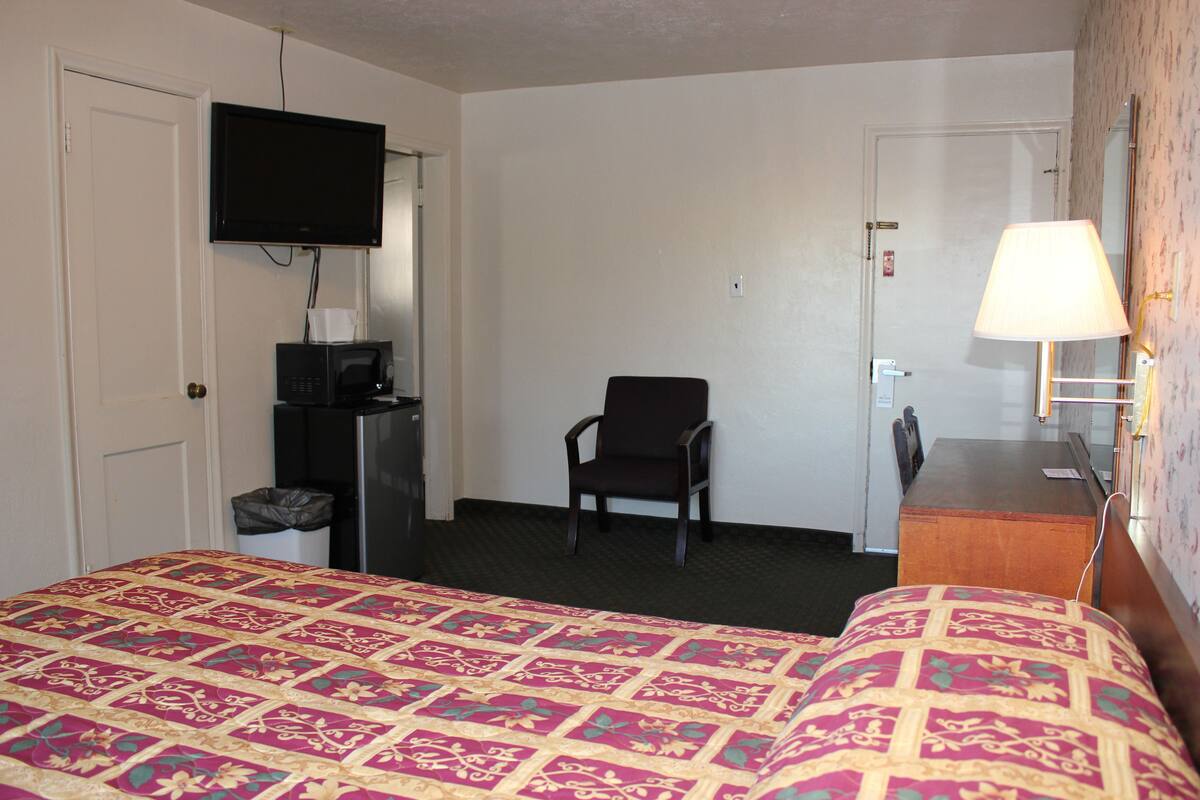 A hotel room with a bed, a TV mounted on the wall, a chair, a desk, and a lamp.