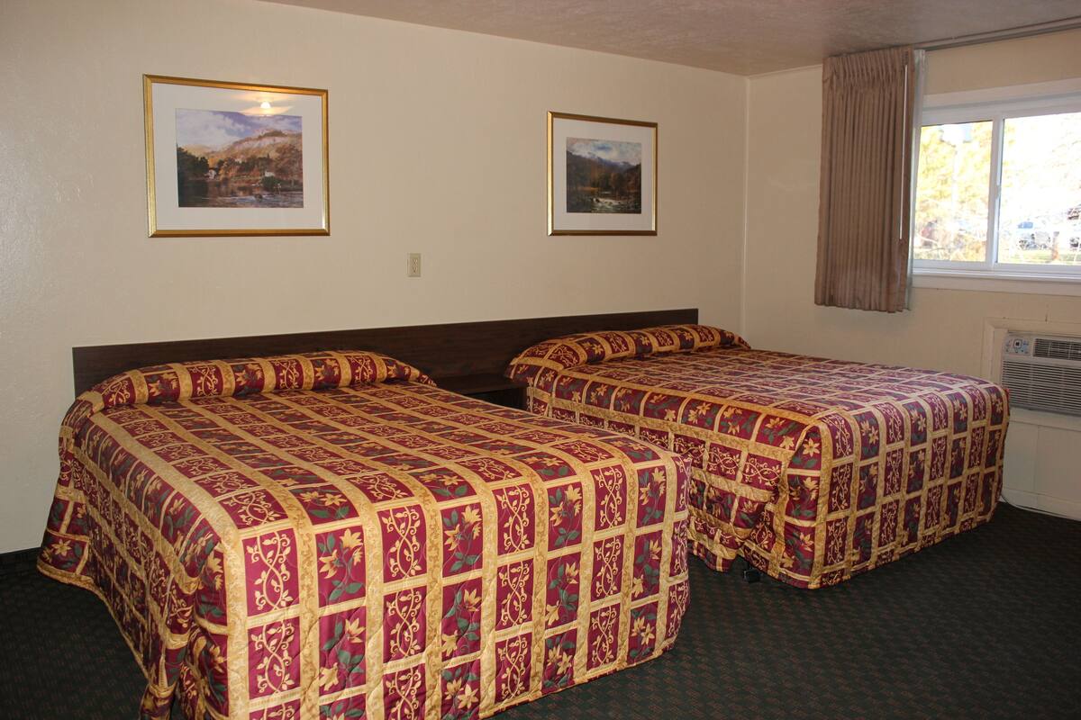 A hotel room with two beds, each with a patterned bedspread, a window with curtains, and two framed pictures on the wall.