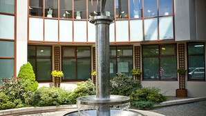 Ein Brunnen mit einer Statue vor einem Gebäude mit großen Fenstern und einem von Ziegelsteinen gepflasterten Platz.