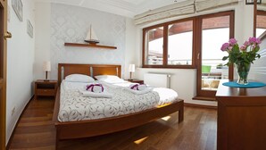 Ein helles Schlafzimmer mit einem hölzernen Bett, einem Nachttisch, einem Beistelltisch, einem Fenster mit Ausblick und einer Blumenvase auf einer Kommode.