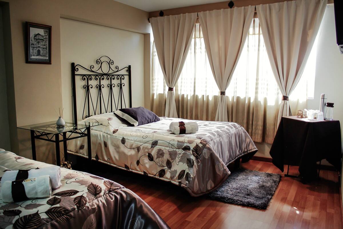 Un dormitorio con dos camas, una mesita de noche con un jarrón, una pequeña mesa con una lámpara y una ventana con cortinas.