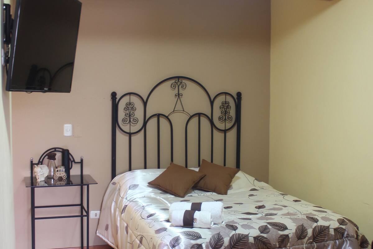 Un dormitorio con cama, una mesita y televisor montado en la pared.