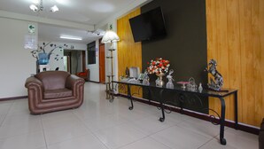 Um hall com um sofá de couro marrom, uma mesa com tampo de vidro e objetos decorativos, uma TV na parede e uma parede de destaque em madeira.