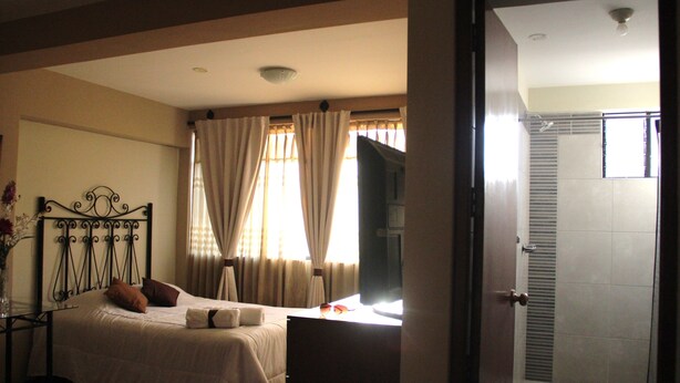 Quarto de hotel com cama, mesa de cabeceira, espelho, cortinas e porta para o banheiro.