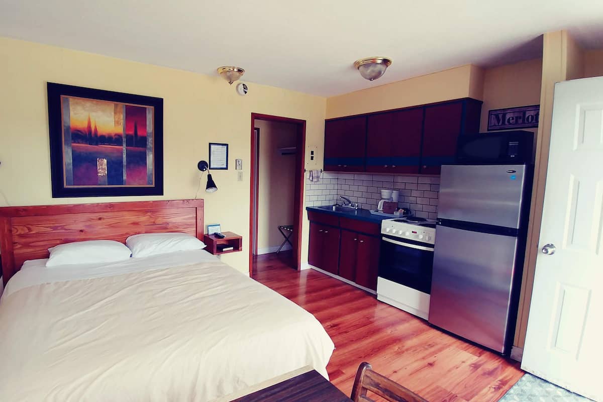 A bedroom with a bed, bedside table, and a painting on the wall. Adjacent is a kitchen with a refrigerator, stove, and cabinets.