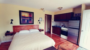 Un dormitorio con cama, mesita de noche y un cuadro en la pared. Junto a ello, una cocina con refrigerador, estufa y alacenas.