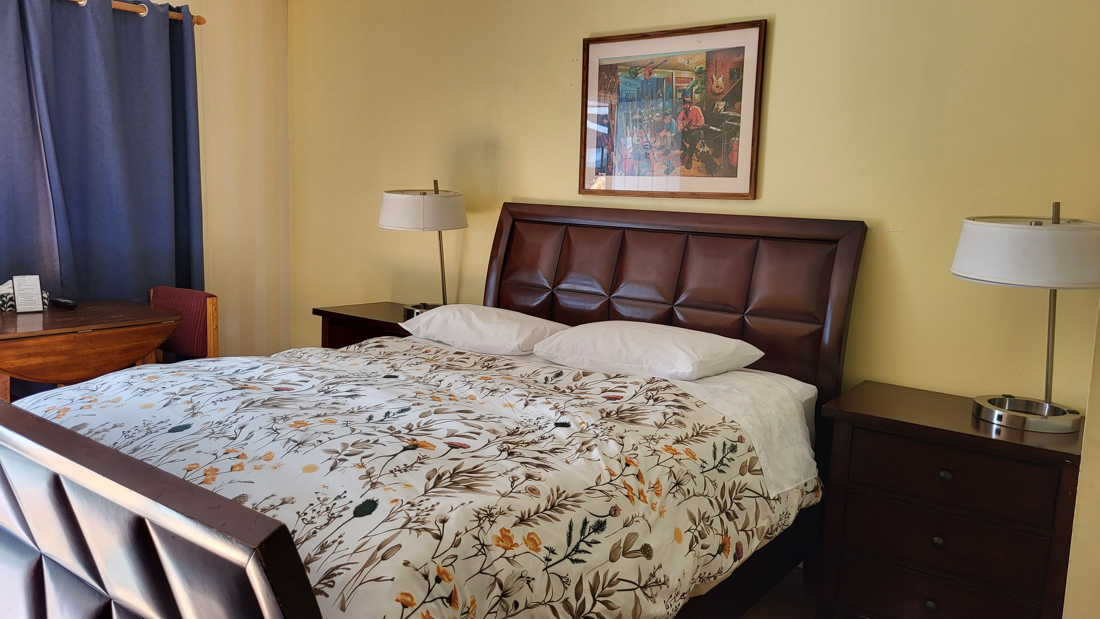 Habitación de hotel con cama, escritorio, silla, dos mesitas de noche con lámparas y un cuadro en la pared.