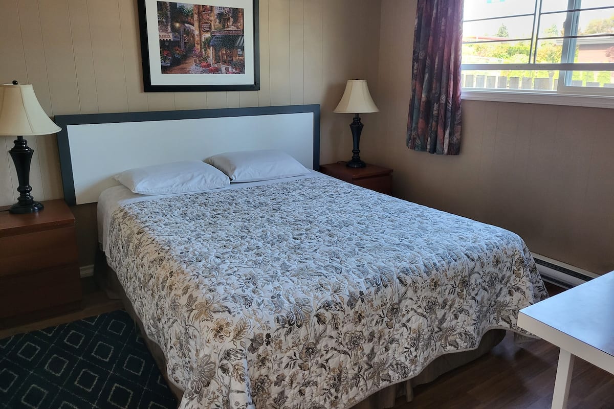 A neatly made bed with a floral bedspread, two bedside lamps, a framed picture on the wall, and a window with curtains.